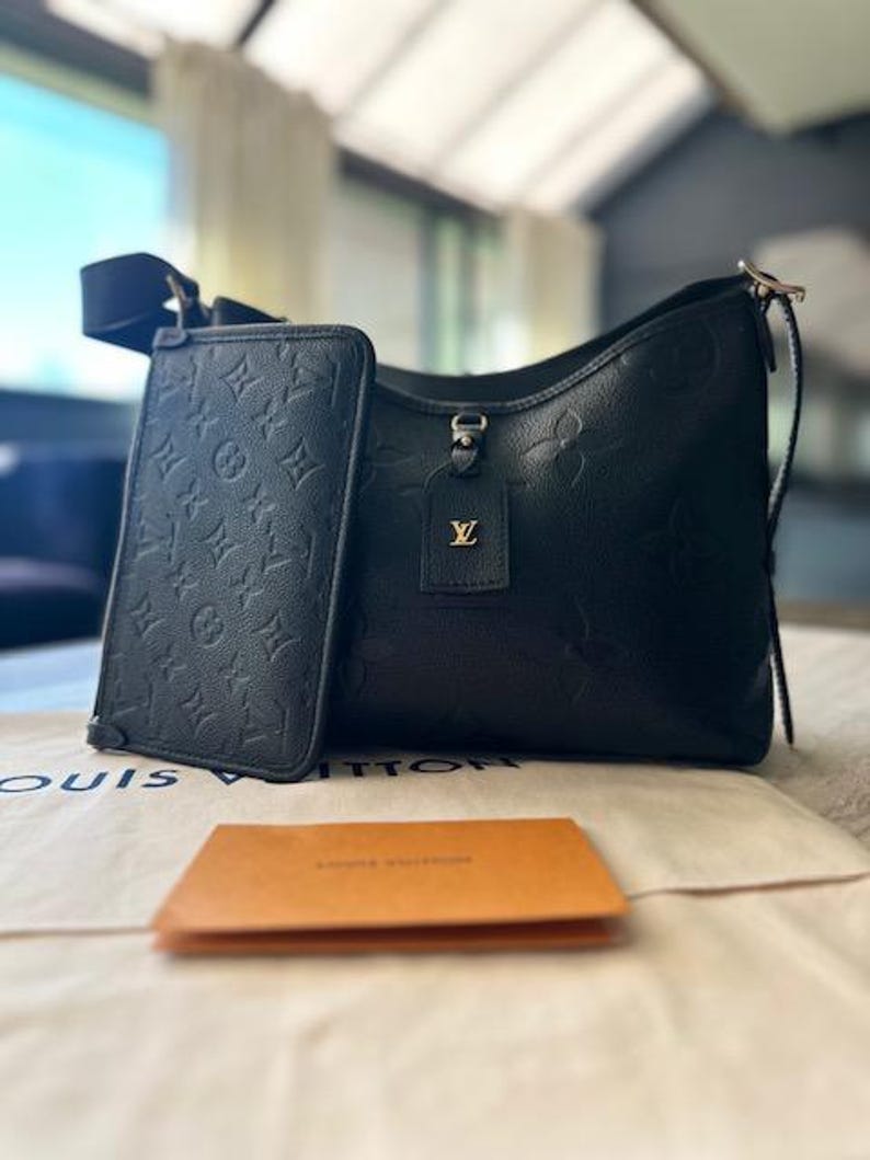 May include: Black leather shoulder bag and matching wallet with embossed monogram pattern. The bag features a top handle and a detachable strap. A small tag with a gold logo hangs from the bag. A small orange card sits in front of the bag.