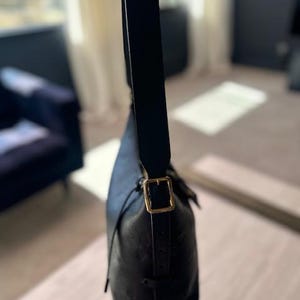 May include: Black leather shoulder bag with a gold-colored buckle. The bag has a long, adjustable strap and a drawstring closure. The leather appears to have a subtle embossed pattern. The bag is shown against a blurred background.