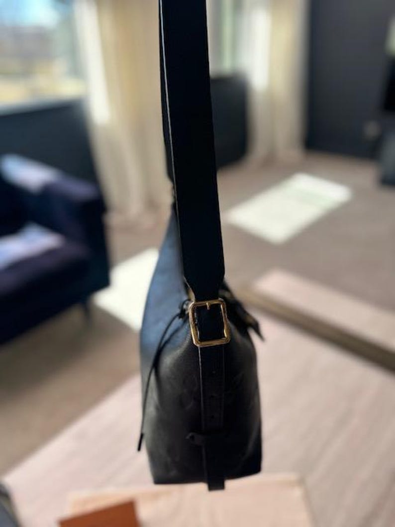 May include: Black leather shoulder bag with a gold-colored buckle. The bag has a long, adjustable strap and a drawstring closure. The leather appears to have a subtle embossed pattern. The bag is shown against a blurred background.