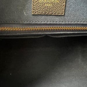 May include: Close-up of a luxury handbag interior. The image shows a dark interior with a gold zipper and a brown leather tag embossed with "LOUIS VUITTON PARIS". The bag's interior is a dark gray color.