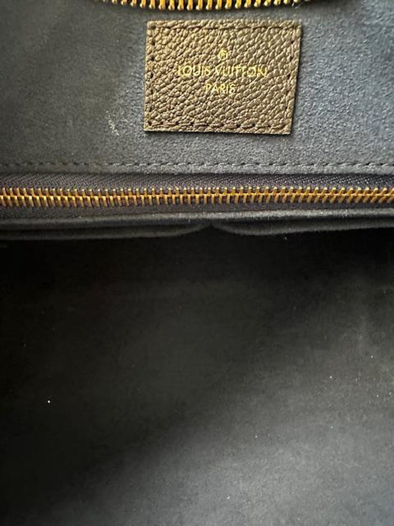 May include: Close-up of a luxury handbag interior. The image shows a dark interior with a gold zipper and a brown leather tag embossed with "LOUIS VUITTON PARIS". The bag's interior is a dark gray color.