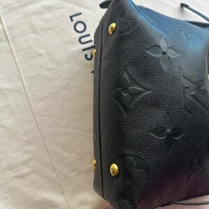 May include: Black leather handbag with embossed floral pattern and gold-tone hardware. The bag is next to a light-colored fabric with the word "LOUIS" printed on it.