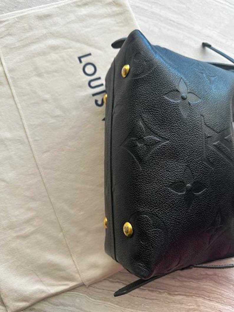 May include: Black leather handbag with embossed floral pattern and gold-tone hardware. The bag is next to a light-colored fabric with the word "LOUIS" printed on it.
