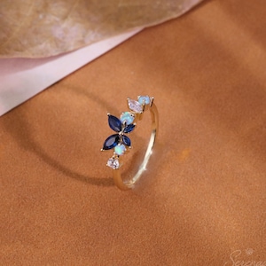 May include: A gold ring featuring a cluster of gemstones arranged in a floral pattern. The design includes marquise-cut dark blue stones, round iridescent opals, and small clear white stones set along a thin, polished gold band.