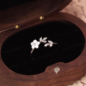Könnte beinhalten: Ein silberfarbener offener Ring mit einer weißen Perlmuttblüte an einem Ende und einer Anordnung aus klaren, marquise-geschliffenen Steinen in Blattform am anderen Ende, präsentiert in einer dunkelbraunen Holzschatulle.
