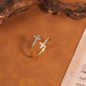 Puede incluir: Un anillo abierto de tono dorado con dos extremos en forma de cruz. Una cruz está adornada con pequeñas piedras azules, mientras que la otra está engastada con piedras blancas transparentes. La banda es lisa y pulida, con un diseño ajustable.