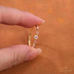 Può includere: Un sottile anello dorato caratterizzato da una delicata disposizione di piccole pietre rosa chiaro taglio marquise e una pietra centrale a forma di fiore bianco. La fascia è sottile e metallica, con un design raffinato e pietre scintillanti.
