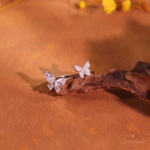 May include: A silver-toned open ring featuring two butterfly-shaped accents, each adorned with clear, sparkling stones. The butterflies are positioned at opposite ends of the band, creating a delicate and symmetrical appearance on the metal.