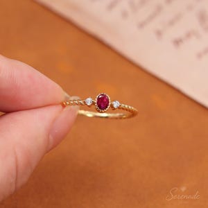 May include: A gold ring featuring a central oval-cut red gemstone flanked by two small round clear stones. The band has a twisted rope-like texture. The setting for the central red stone is scalloped, and the ring is held against a blurred background.