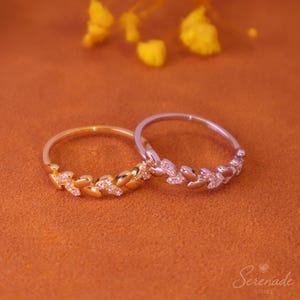 May include: Two rings featuring a delicate leaf-shaped band design. One ring is gold-toned and the other is silver-toned, both accented with small, clear, sparkling stones embedded along the leaf motifs. The rings are positioned side by side.