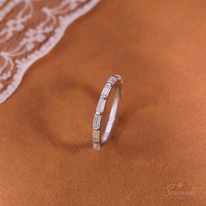 May include: A silver-toned band ring featuring a series of clear, rectangular baguette-cut stones set in a row along the outer surface. The ring has a polished metallic finish and a slim, uniform profile, showcasing a clean and structured design.