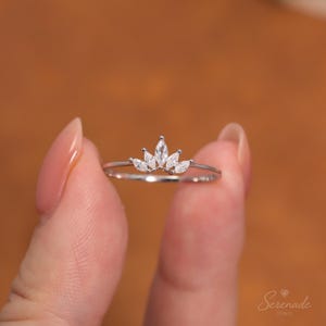 May include: A silver-toned ring featuring a delicate crown-shaped design adorned with five marquise-cut clear stones. The band is thin and polished, showcasing a symmetrical arrangement of stones that taper in size from the center outward.