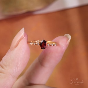May include: A gold ring featuring a central oval-cut deep red garnet gemstone. The band is accented with three small clear stones on each side of the centre setting. The text Serenade Stones is visible in the bottom right corner of the image.