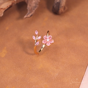May include: An open-style gold ring featuring a floral design on one end with five pink marquise-cut stones forming petals around a central clear stone, and a branch-like motif on the other end with three small pink marquise-cut leaves.