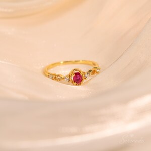 May include: A gold ring featuring a central oval-cut red gemstone surrounded by a beaded metal frame, flanked by small clear stones on each side of the band. The band has a delicate, textured design with intricate metalwork details.