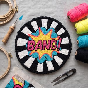 May include: A circular punch needle embroidery piece featuring a comic book style explosion graphic with the word BANG! in pink letters, surrounded by a yellow and blue burst on a black and white striped background, with yarn and tools.