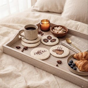 May include: A light-colored wooden serving tray holds a speckled ceramic mug, a lit candle in a glass jar, a wooden bowl of sugar cubes, and three round, textured coasters featuring coffee-themed designs. A plate with croissants and blueberries sits nearby.