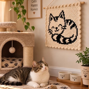 May include: A square cream-coloured crochet wall hanging featuring a black cat graphic, suspended from a wooden dowel with a rope. The piece has a scalloped border and displays a stylised cat design with stripes, whiskers, and a smiling face.