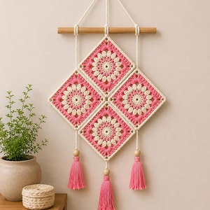 May include: A wall hanging featuring three diamond-shaped crochet squares in pink and cream, arranged in a vertical pattern. The squares are suspended from a horizontal wooden dowel by white cords, with three pink yarn tassels hanging from the bottom.