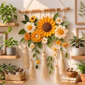 Op de afbeelding: Een wandhanger met gehaakte zonnebloemen, madeliefjes en goudsbloemen in geel, wit en oranje met groene bladeren. Het bloemstuk hangt aan een houten stok met crèmekleurige koorden, versierd met houten kralen en kwastjes.