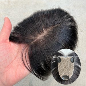 May include: A hair topper featuring straight, dark black hair with a visible scalp-like base at the crown. The underside of the hairpiece shows a beige, mesh-like foundation with three small metal pressure-sensitive clips attached for secure placement.