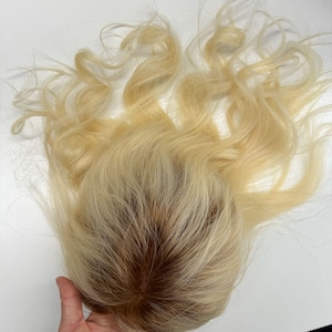 May include: A blonde hairpiece with wavy, light-colored strands. The hairpiece has a darker, brown-toned base, and the hair is styled with loose curls. The hairpiece is likely a wig or hair topper.