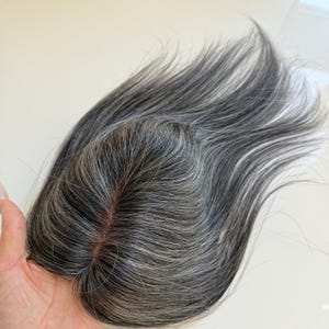 May include: A hair topper featuring a blend of dark charcoal and silver-grey strands. The piece has a central part with a visible scalp-colored base, showing straight, smooth hair fibers that flow outward from the crown in a natural layered style.