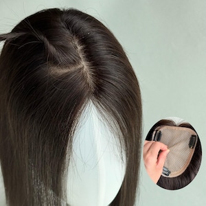 Puede incluir: Un postizo de cabello castaño oscuro con una base de monofilamento y una raya de aspecto natural. La imagen insertada muestra la parte inferior de la pieza, que incluye una base de malla beige y clips metálicos para una fijación segura.