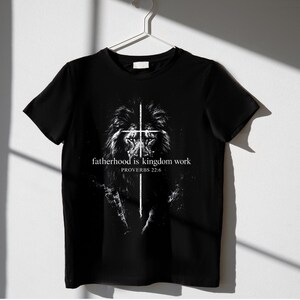 May include: Black t-shirt featuring a detailed white illustration of a lion's face with a cross superimposed over it. The text "fatherhood is kingdom work" and "PROVERBS 22:6" are printed in white below the lion image.