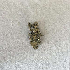 Puede incluir: Un broche de metal color latón que presenta un relieve detallado de una figura con armadura que sostiene un arma de asta. La figura está colocada sobre una base decorativa en forma de escudo curvo con un diseño ornamentado y en espiral.