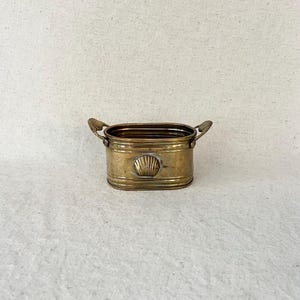 May include: An oval brass planter featuring a polished metallic finish, two small side handles, and a decorative embossed seashell motif centered on the front. The vessel has a horizontal ribbed texture along the upper and lower rim sections.