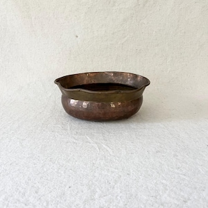 May include: A round, hammered copper bowl featuring a smooth, wide brass rim. The bowl has a deep, reddish-brown metallic finish with visible indentations from the hammering process along the exterior surface and a dark, reflective interior.