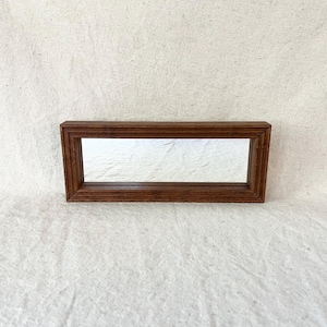 May include: A rectangular, wooden framed mirror with a dark brown finish. The mirror's reflective surface is centered within the frame. The frame has a simple, beveled edge design. The mirror is a decorative item.