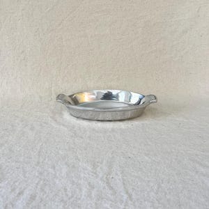 May include: Oval, silver serving dish with two small handles. The dish has a reflective surface and is empty. The background is a neutral, off-white fabric. The dish is likely made of metal.