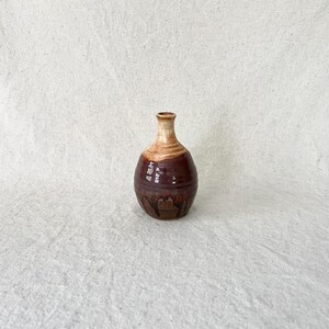 Può includere: Un piccolo vaso in ceramica con corpo arrotondato e collo stretto. La parte superiore presenta una smaltatura beige chiaro, mentre la parte inferiore è rifinita con uno smalto marrone scuro lucido con sottili motivi a goccia verticali.