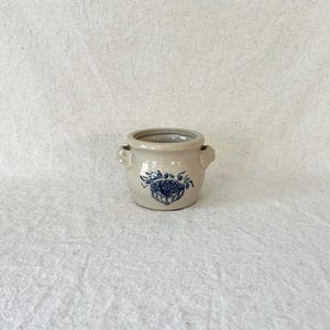 May include: A small, beige ceramic pot with two small handles and a blue floral design. The pot has a round shape and is likely used for decorative purposes or small plants. The blue design is centered on the front of the pot.