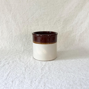 Puede incluir: Un recipiente cilíndrico de cerámica con un diseño de dos tonos. La parte superior es marrón oscuro, la sección media es marrón claro y la base es de color blanco crema. El recipiente tiene un diseño simple y clásico.