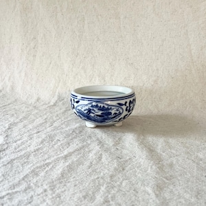May include: A small, round, blue and white ceramic bowl with a detailed landscape design. The bowl has a scalloped rim and sits on three small feet. The interior of the bowl appears to contain a small amount of liquid.