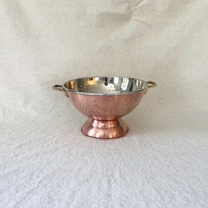 Puede incluir: Un colador redondo con exterior de cobre pulido e interior de metal plateado. El cuenco está perforado con pequeños orificios de drenaje, se asienta sobre una base circular y cuenta con dos pequeñas asas metálicas en los lados.