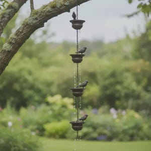 May include: A decorative rain chain with four tiers of bronze-colored bowls, each adorned with a small bird figurine. The chain hangs from a tree branch, with water flowing down the chain. The background is a blurred green garden.