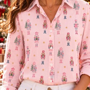 May include: A light pink long-sleeve button-down shirt featuring a repeating pattern of nutcracker figures in shades of pink, blue, and white, interspersed with small gold stars. The shirt has a classic collar and white buttons.