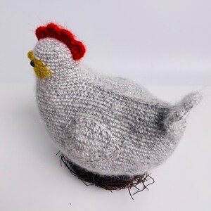 May include: A handmade knitted hen figurine in shades of gray, with a bright red comb and yellow beak. The hen is perched on a woven nest made of brown twigs, creating a rustic and charming decorative piece.