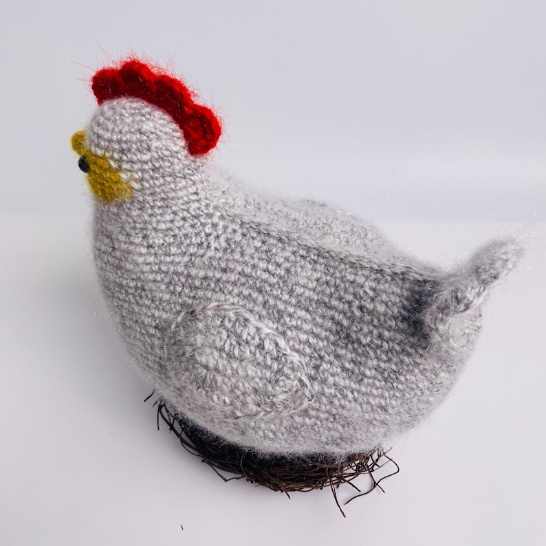 May include: A handmade knitted hen figurine in shades of gray, with a bright red comb and yellow beak. The hen is perched on a woven nest made of brown twigs, creating a rustic and charming decorative piece.
