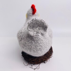 May include: A handmade gray knitted chicken figurine with a red comb and yellow accents, perched on a brown twig nest. The chicken is viewed from the back, showcasing its textured body and wings.