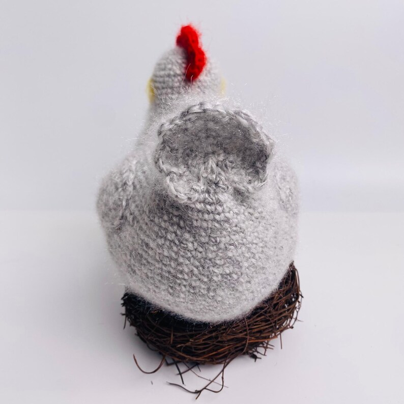 May include: A handmade gray knitted chicken figurine with a red comb and yellow accents, perched on a brown twig nest. The chicken is viewed from the back, showcasing its textured body and wings.