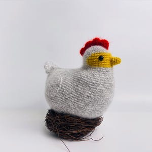 May include: A handmade, gray knitted chicken with a red comb, yellow beak, and black eye, perched on a small, brown twig nest. The chicken is a whimsical decorative item, perfect for farmhouse or country-themed decor.