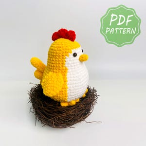 Puede incluir: Un juguete de peluche de pollo tejido a ganchillo en amarillo y blanco con una cresta roja, ojos de cuentas negras y un pequeño pico amarillo, sentado dentro de un nido de ramitas marrones. Una insignia verde festoneada en la esquina superior derecha contiene el texto PDF PATTERN.