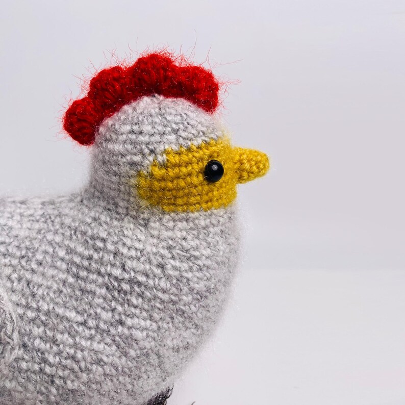 May include: A handmade knitted chicken toy. The chicken has a gray body, a red comb, a yellow beak, and a black eye. The toy is made of soft yarn and is designed to look like a cartoon chicken.