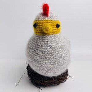 May include: A handmade, gray crocheted chicken figurine with a yellow face and red comb, perched on a brown twig nest. The chicken has black eyes and a fluffy texture, creating a whimsical, decorative piece.