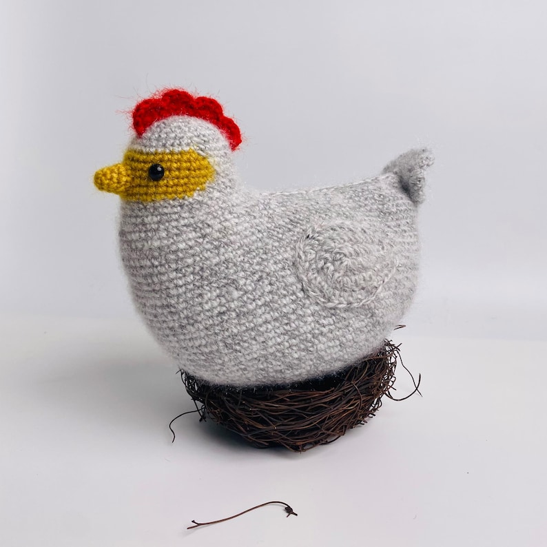May include: A handmade crochet chicken in shades of gray, yellow, and red, perched on a small brown nest. The chicken has a red comb, yellow beak, and a gray body. A charming, decorative item.